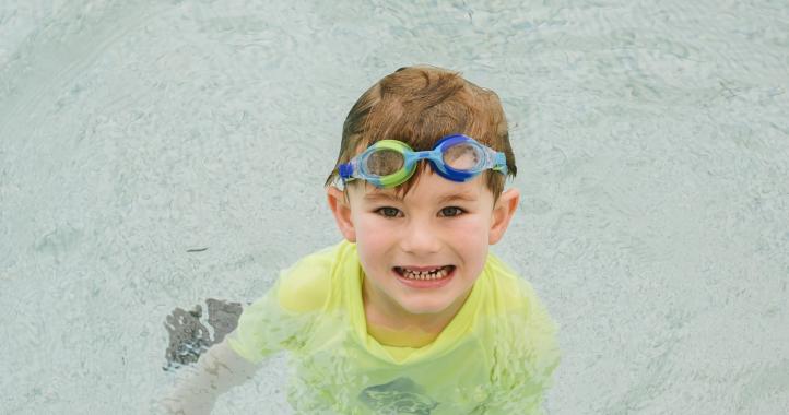 Boy with googles and yellow rash vest smiling at camera in a pool